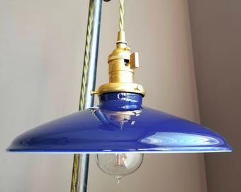 Puede incluir: Una lámpara colgante azul con un accesorio de latón y una bombilla de estilo vintage. La luz está suspendida de un sistema de poleas de madera con un cable trenzado verde y amarillo. La lámpara tiene una pantalla de esmalte azul brillante.