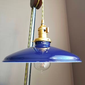 Puede incluir: Una lámpara colgante azul con un accesorio de latón y una bombilla de estilo vintage. La luz está suspendida de un sistema de poleas de madera con un cable trenzado verde y amarillo. La lámpara tiene una pantalla de esmalte azul brillante.