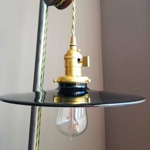 Puede incluir: Una lámpara colgante de metal negro con una toma de corriente dorada y una bombilla de vidrio transparente. La lámpara está suspendida de un cordón trenzado verde.