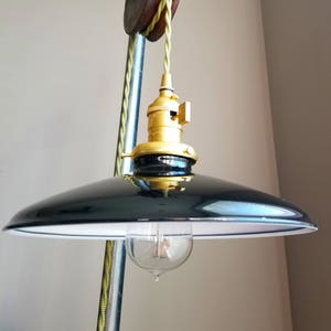 Puede incluir: Una pantalla de lámpara de metal negro con un interior blanco cuelga de un sistema de polea y cordón de color dorado. El cordón es verde y la polea está unida a un tubo de metal.