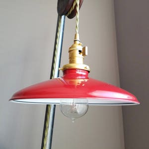 Puede incluir: Una lámpara colgante de metal roja con un interior blanco y una toma de corriente dorada. La luz está suspendida de una cuerda verde y un sistema de poleas vintage.