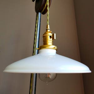 May include: A white glass pendant light fixture with a gold metal socket and a white cord hanging from a vintage wooden pulley. The light is suspended from a metal pipe.