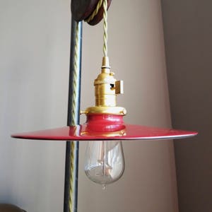 Puede incluir: Una pantalla de lámpara de metal roja con ribete dorado cuelga de un sistema de poleas vintage con un cordón trenzado verde. Se ve una bombilla de vidrio transparente.