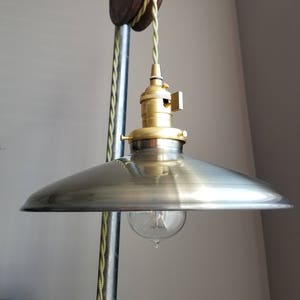 Puede incluir: Una pantalla de lámpara de metal plateado con un casquillo y un cable de metal dorado que cuelgan de un sistema de poleas vintage. El cable es de color verde claro.