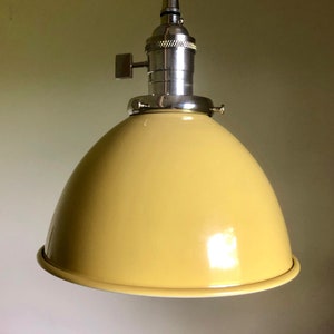 Puede incluir: Una lámpara colgante de metal amarilla con un casquillo plateado y un interruptor de encendido/apagado simple. La luz tiene una pantalla en forma de cúpula.
