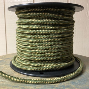 Puede incluir: Una bobina negra que contiene un cordón trenzado verde oliva y beige. El cordón está enrollado firmemente, mostrando un patrón texturizado. El cordón es adecuado para manualidades y proyectos de diseño. El diámetro del cordón es de aproximadamente 0,6 cm.