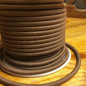 Puede incluir: Un carrete de cordón elástico marrón, bien enrollado y apoyado sobre una base blanca. El cordón es de un color marrón rico y terroso, y la textura parece suave y ligeramente borrosa. El cordón probablemente se utiliza para proyectos de artesanía o costura.