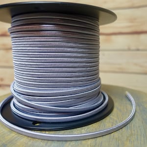 May include: A spool of silver braided cord, wound around a black plastic spool. The cord is smooth and shiny, with a subtle texture.