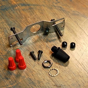 Może przedstawiać: Metalowa skrzynka elektryczna z dwoma śrubami, trzema czerwonymi złączami przewodów, dwoma czarnymi końcówkami przewodów, dwoma małymi czarnymi śrubami, dwoma większymi czarnymi śrubami, czarnym złączem przewodów, metalową podkładką i metalowym pierścieniem.