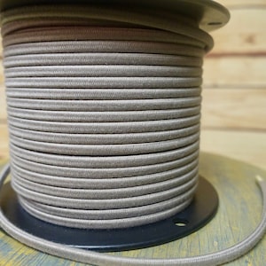 Puede incluir: Una bobina de cordón de tela beige enrollada alrededor de una bobina de plástico negro. El cordón es liso y tiene una textura sutil.