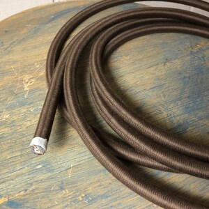 May include: A close-up of brown braided cord with a white and silver end cap. The cord is coiled on a wooden surface.