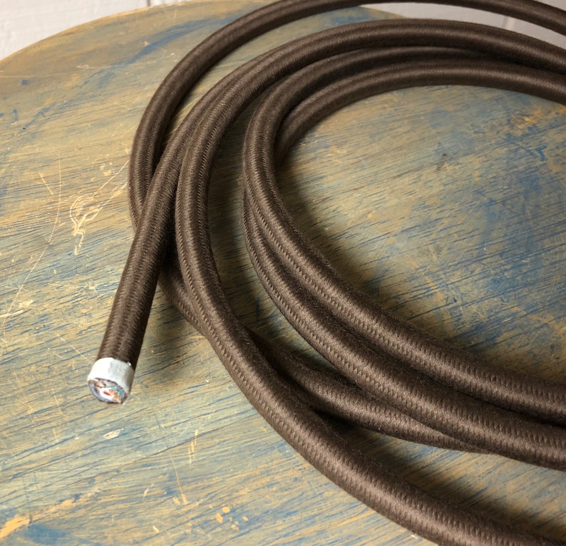 May include: A close-up of brown braided cord with a white and silver end cap. The cord is coiled on a wooden surface.