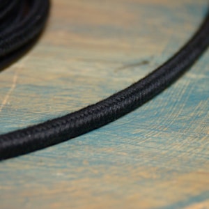 May include: Close-up of black electrical cord with a braided fabric covering. The cord is lying on a blue and brown wooden surface.