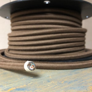 May include: A spool of brown cloth-covered electrical cord. The cord is wound tightly around the spool and the end of the cord is visible, showing the white insulation and the wires inside.