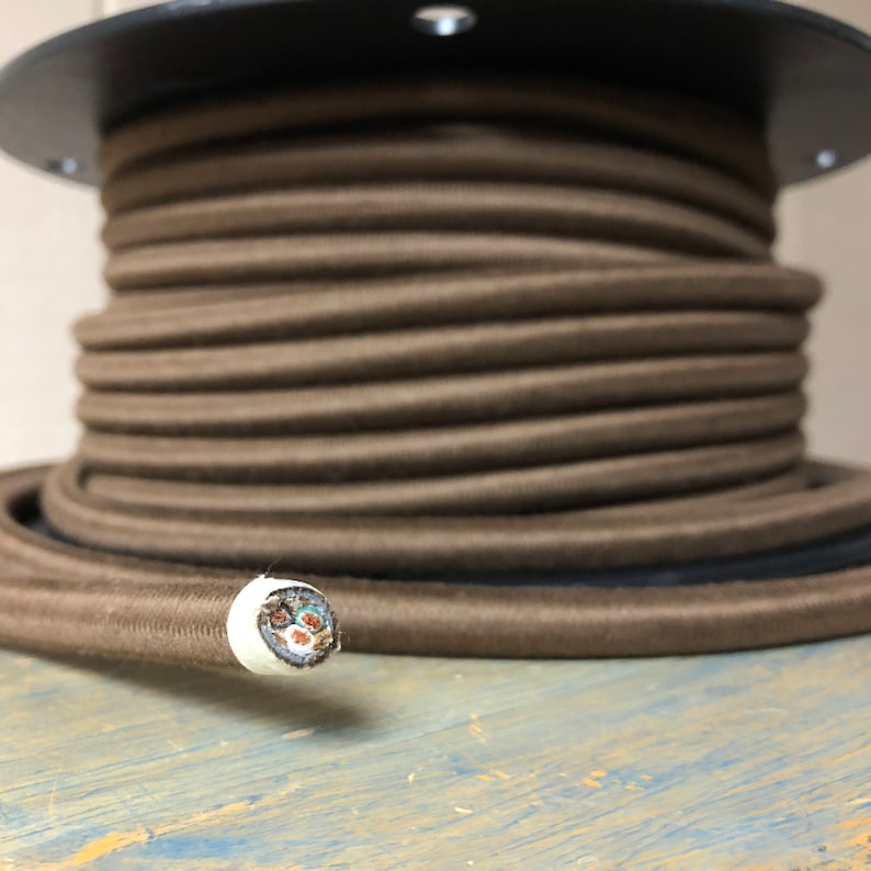 May include: A spool of brown cloth-covered electrical cord. The cord is wound tightly around the spool and the end of the cord is visible, showing the white insulation and the wires inside.