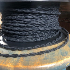 May include: A spool of black braided electrical cord. The cord is wound tightly around the spool and is ready to be used for electrical projects.