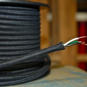 Peut inclure: Une bobine de fil électrique recouvert de tissu noir avec trois fils exposés à l'extrémité. Les fils sont blanc, vert et cuivre.