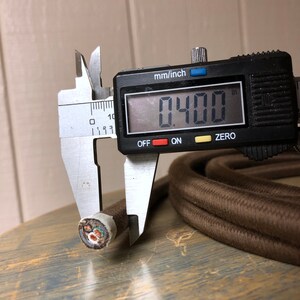 May include: Close-up of a brown fabric-covered electrical cord being measured with a digital caliper. The caliper reads 0.400 inches. The cord has three wires inside.