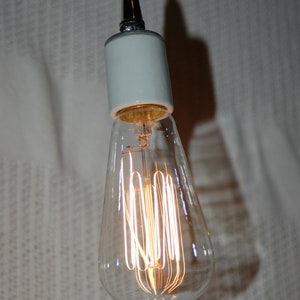 May include: A white ceramic light fixture with a black cord hanging from the ceiling. The light fixture has a clear glass bulb with a filament that is lit.