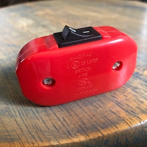 以下が含まれることがあります： 黒いトグルスイッチが付いた赤いプラスチック製のオン/オフスイッチ。スイッチには、「120VAC」、「US Listed」、「Switch」、「1 YR」、「WE」、「CHINA-W」、「CAT NO. 1445」と表示されています。