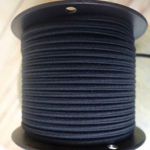 Puede incluir: Una bobina de cordón elástico negro enrollado alrededor de una bobina de plástico negro. El cordón elástico está enrollado apretado y parece ser de buena calidad.