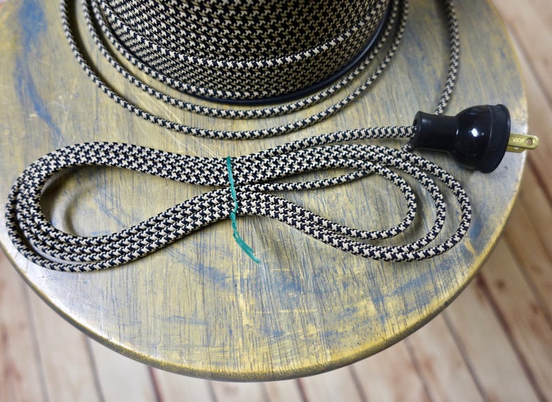 May include: Black and white houndstooth patterned electrical cord with a black plug. A spool of matching cord is visible in the background. The cord is looped and secured with a green tie, suggesting a craft or DIY project.
