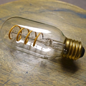 May include: A clear, oblong-shaped light bulb with a gold base and a spiral filament. The filament is a warm yellow color, and the bulb is resting on a wooden surface. The bulb is designed for decorative lighting.