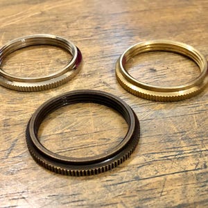 Op de afbeelding: Drie metalen ringen met een schroefdraad aan de binnenkant en een geribbelde buitenkant. Eén ring is zilverkleurig, één is goudkleurig en de derde is donkerbrons. De ringen zijn gerangschikt op een houten oppervlak.