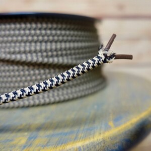 May include: Close-up of a decorative electrical cord with a houndstooth pattern in navy blue and white. The cord is unwound, revealing two brown wires. A spool of the same cord is in the background, resting on a blue and yellow surface.
