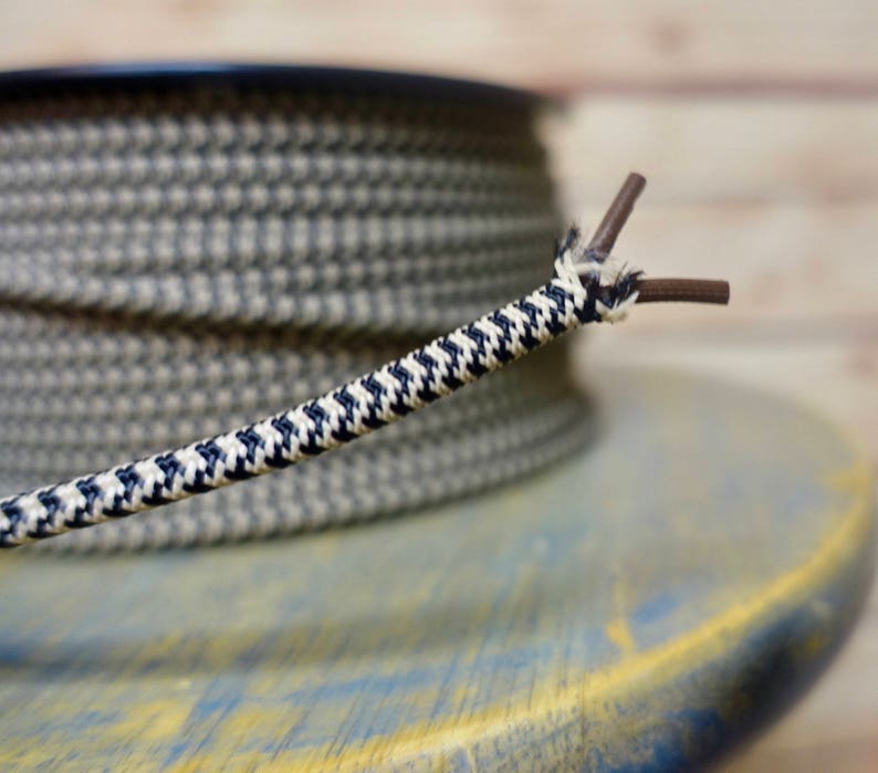 May include: Close-up of a decorative electrical cord with a houndstooth pattern in navy blue and white. The cord is unwound, revealing two brown wires. A spool of the same cord is in the background, resting on a blue and yellow surface.