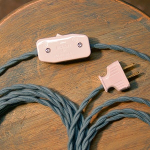 May include: A close-up of a teal blue fabric-covered electrical cord with a white on/off switch and a white plug. The cord is coiled and ready to be used.