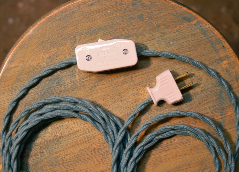 May include: A close-up of a teal blue fabric-covered electrical cord with a white on/off switch and a white plug. The cord is coiled and ready to be used.