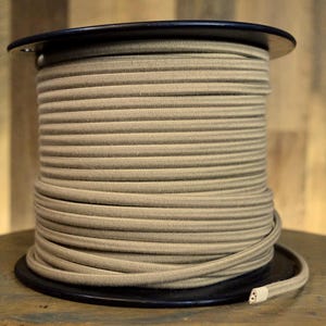 Puede incluir: Un carrete de madera negro que contiene una gran cantidad de cordón elástico beige. El cordón está cuidadosamente enrollado, formando una forma cilíndrica. El extremo del cordón es visible, con una punta blanca. El fondo es una superficie de madera.