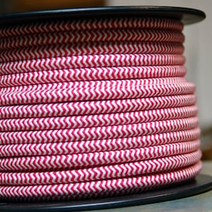 Puede incluir: Un carrete de cable eléctrico rojo y blanco con estampado de chevrón. El cable está enrollado firmemente alrededor de un carrete de plástico negro. El patrón es un diseño en zigzag repetitivo. El cable probablemente se utiliza para iluminación u otros proyectos eléctricos.