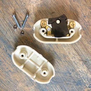 Puede incluir: Dos tapas de tomacorriente de pl&aacute;stico beige con un interruptor de palanca negro en posici&oacute;n ON. El interruptor tiene las palabras ON y OFF impresas. Hay dos tornillos plateados y dos tuercas plateadas sobre una superficie de madera.