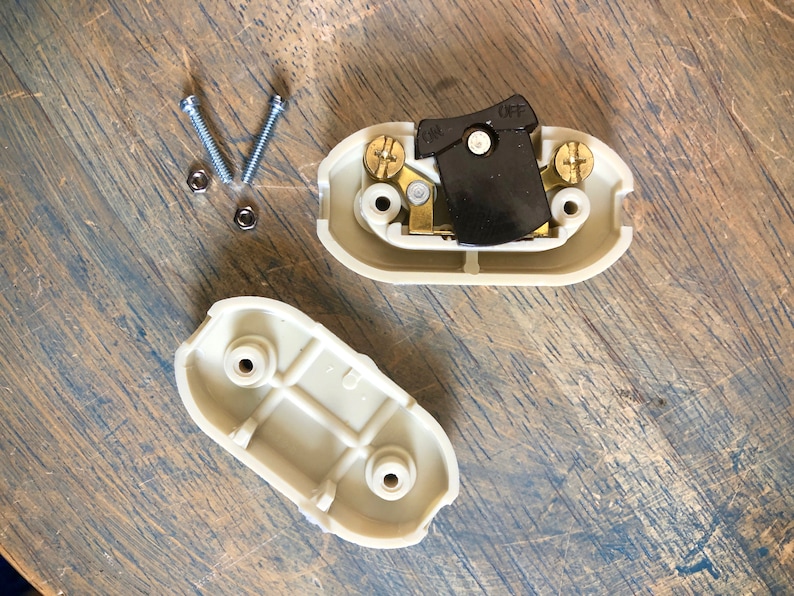 Puede incluir: Dos tapas de tomacorriente de pl&aacute;stico beige con un interruptor de palanca negro en posici&oacute;n ON. El interruptor tiene las palabras ON y OFF impresas. Hay dos tornillos plateados y dos tuercas plateadas sobre una superficie de madera.