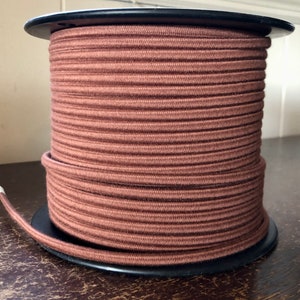 Puede incluir: Una bobina de cordón de tela marrón, enrollada firmemente alrededor de una bobina de plástico negro. El cordón es liso y tiene una textura sutil.