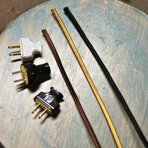 May include: Three different types of electrical cords, including brown, black, and gold, are shown on a light blue wooden surface.  There are also three electrical plugs, one white, one black, and one brown.