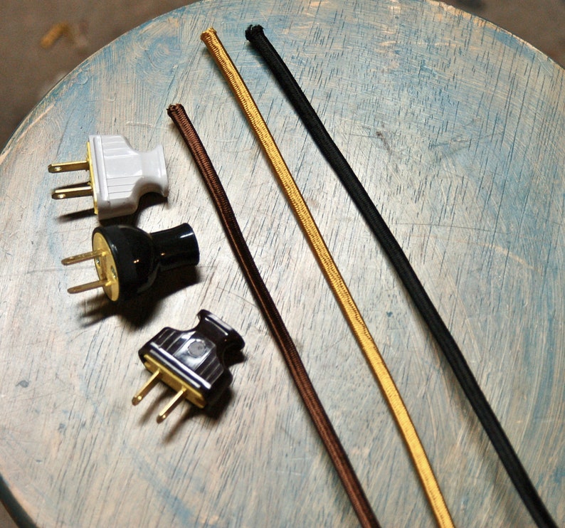 May include: Three different types of electrical cords, including brown, black, and gold, are shown on a light blue wooden surface.  There are also three electrical plugs, one white, one black, and one brown.
