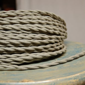 May include: A close-up of a spool of thick, braided, light gray electrical cord. The cord is coiled neatly and is sitting on a light blue wooden surface.