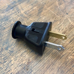 May include: A black electrical plug with two flat, silver prongs and a gold-coloured grounding pin. The plug has a cylindrical end and a rectangular body with a textured design. It is designed for electrical devices.