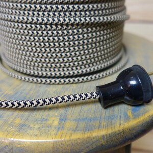 May include: A spool of black and cream-colored fabric-covered electrical wire with a matching plug. The wire has a patterned design and is coiled on a wooden spool. The plug is black with two prongs.