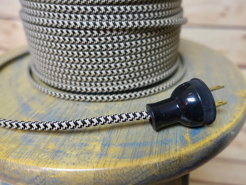 May include: A spool of black and cream-colored fabric-covered electrical wire with a matching plug. The wire has a patterned design and is coiled on a wooden spool. The plug is black with two prongs.