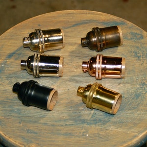 May include: Six different light bulb sockets in various finishes, including brass, copper, nickel, and black. The sockets are arranged on a wooden surface.