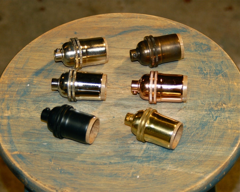 May include: Six different light bulb sockets in various finishes, including brass, copper, nickel, and black. The sockets are arranged on a wooden surface.
