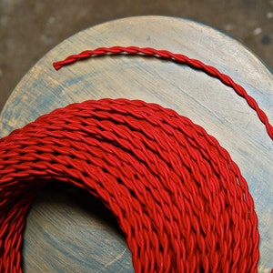 Puede incluir: Una bobina de alambre eléctrico rojo trenzado. El alambre está enrollado firmemente alrededor de sí mismo, creando una bobina roja gruesa.
