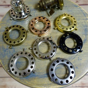 Puede incluir: Surtido de ocho anillos de casquillo de lámpara de metal en varios acabados, incluyendo níquel, latón, negro y cobre. Cada anillo tiene un diseño circular con múltiples agujeros alrededor del borde exterior.