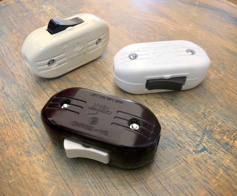 Puede incluir: Tres interruptores de luz vintage, uno blanco, uno negro y uno crema. Cada interruptor tiene una palanca de conmutaci&oacute;n y est&aacute; etiquetado con el nombre de la marca "Cooper" y el texto "125V-15A".
