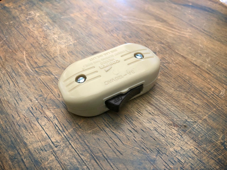 Puede incluir: Un interruptor de luz de pl&aacute;stico beige con un interruptor de palanca marr&oacute;n. El interruptor est&aacute; etiquetado como "Cooper" y "5A-120VAC 1000 Watt Max. 1/2 HP 125 VAC. El interruptor est&aacute; sobre una superficie de madera.