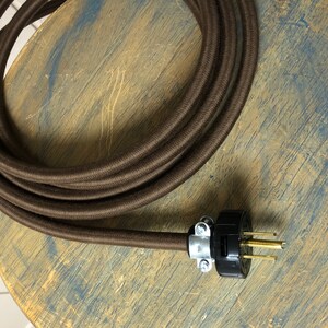 May include: Brown cloth-covered electrical cord with a black and silver plug. The cord is coiled on a wooden surface.